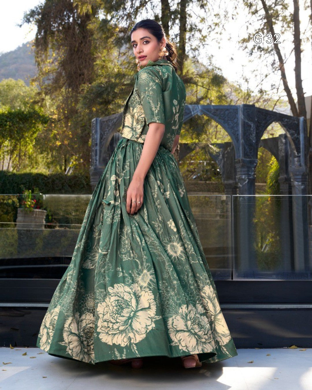 A woman wearing a green and beige floral print maxi dress, standing outdoors in front of a modern architectural background with trees.