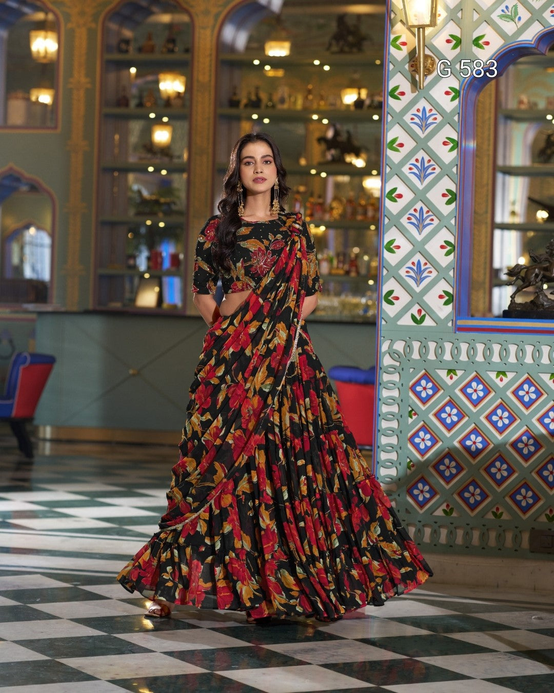 A woman models a black floral printed maxi dress with a matching printed blouse in an ornate, traditional Indian interior with patterned walls and a checkered floor.