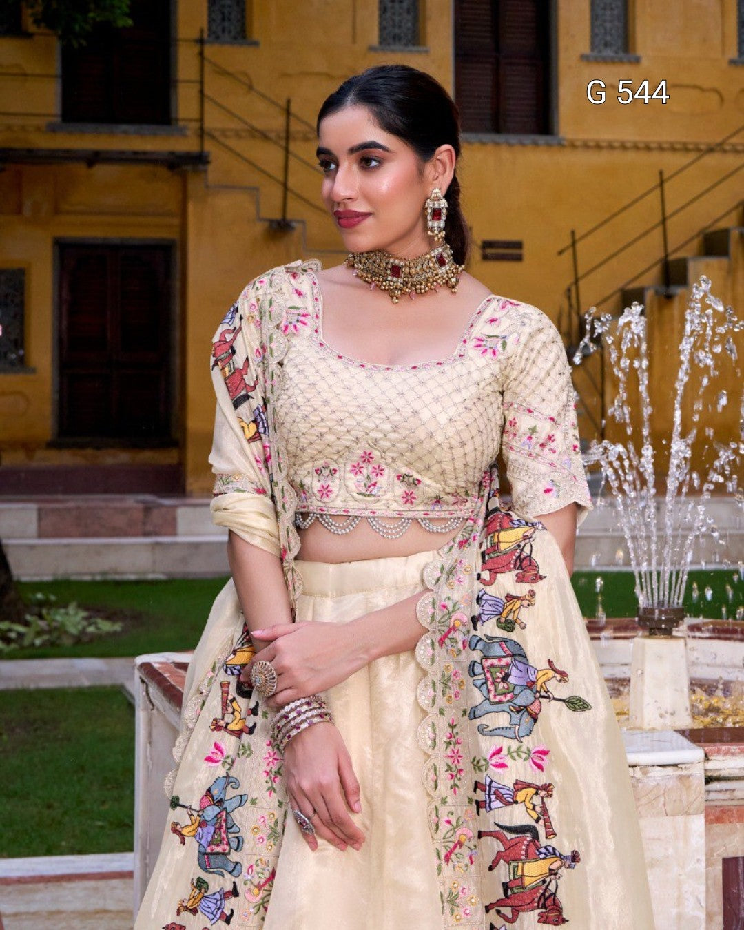 A woman models a cream-colored traditional Indian ethnic outfit with floral embroidery and colorful printed motifs on the dupatta, set against a yellow building background.