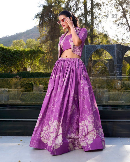 A woman modeling a purple two-piece traditional outfit with a matching flared skirt and cropped top, featuring a light floral print. She is standing outdoors in front of a glass railing and greenery.