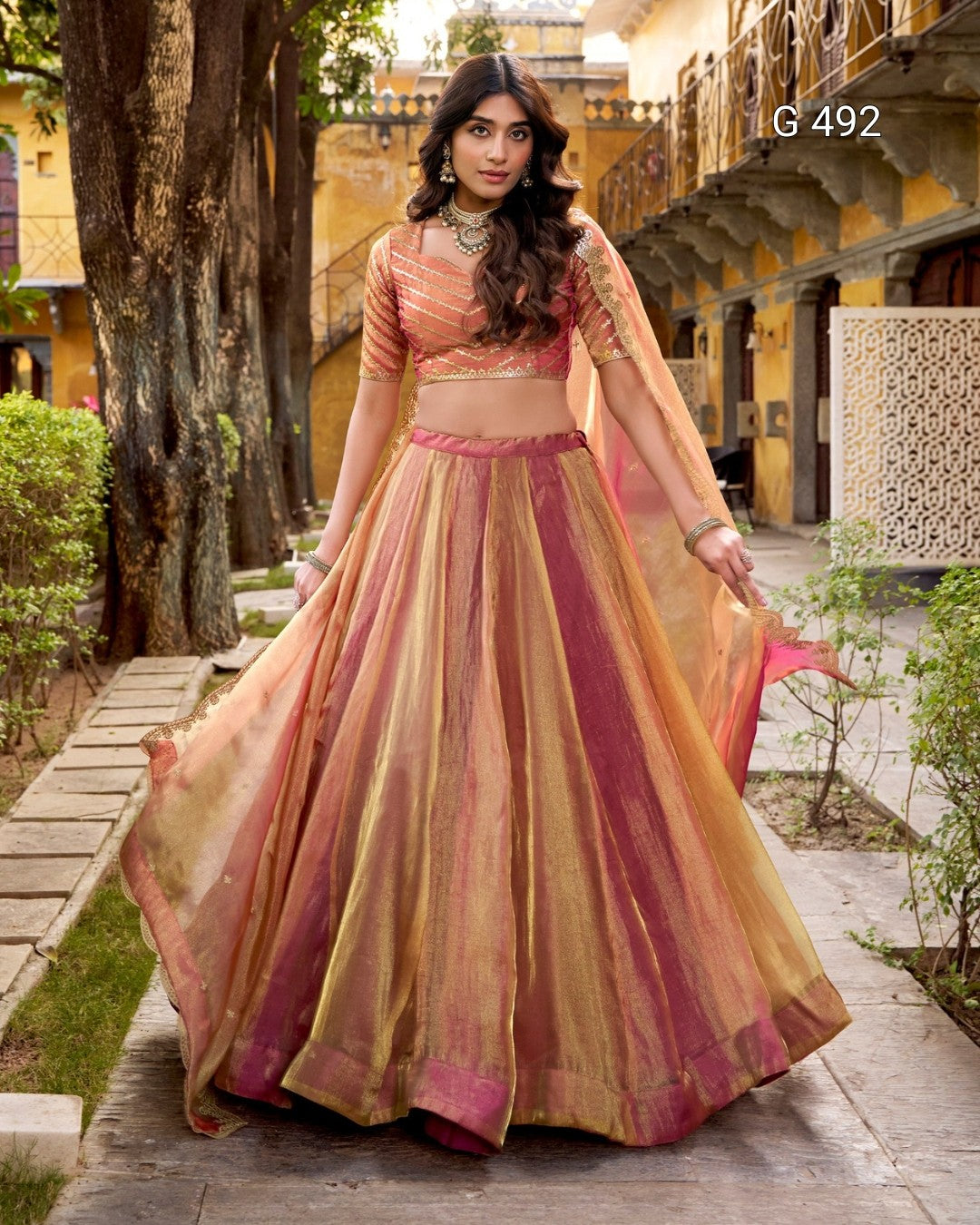 A woman models a traditional Indian lehenga choli set in an outdoor courtyard. The outfit features a sheer peach and gold embroidered blouse with a matching flared skirt in peach, gold, and deep pink, with a sheer dupatta draped over her shoulder.