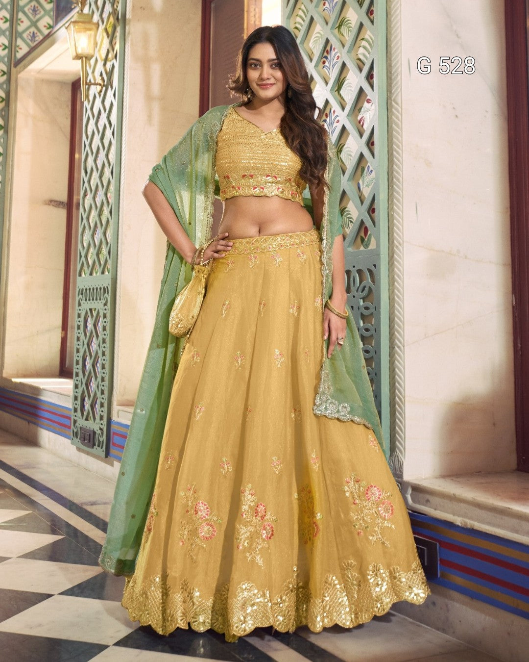 A woman models a traditional Indian ethnic wear set consisting of a mustard yellow embroidered lehenga skirt, matching crop top, and a light green dupatta, standing in an ornate architectural corridor.