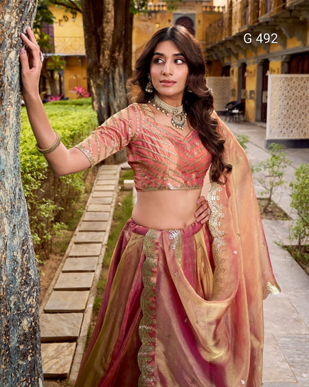 A woman models a traditional Indian salwar kameez outfit in a garden setting. The outfit consists of a peach-pink and gold embroidered crop top, a matching pink and gold skirt, and a sheer peach dupatta.