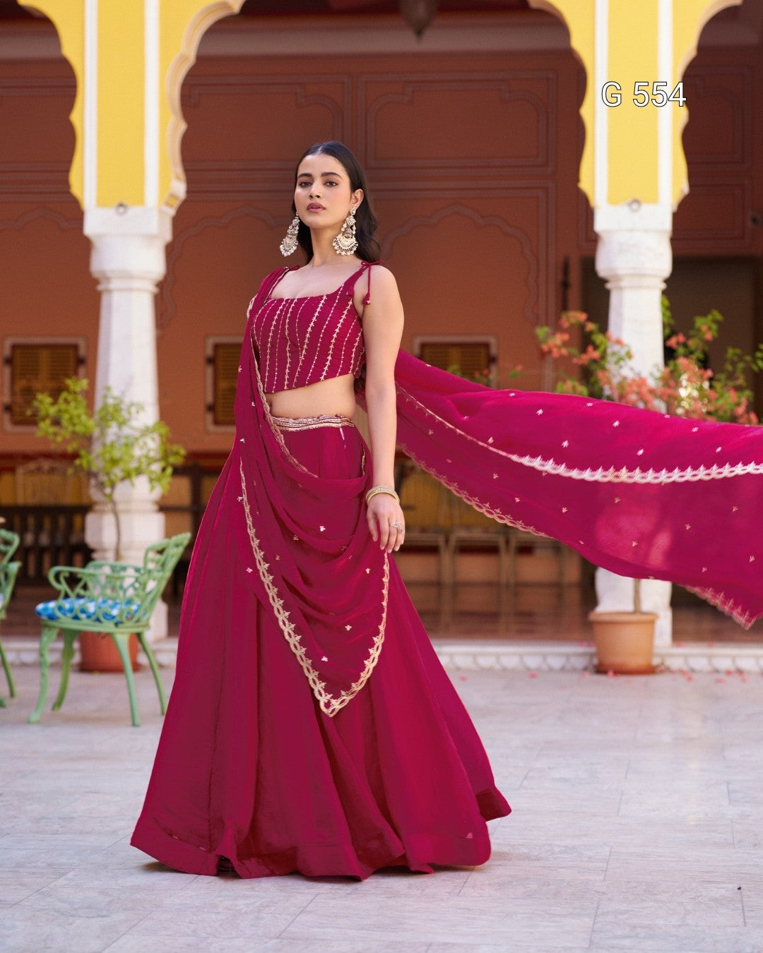 A woman models a deep magenta lehenga choli set with gold embroidery, standing in front of a traditional courtyard with yellow arches. The dupatta is spread out to the side, showing its gold border and small gold embellishments.