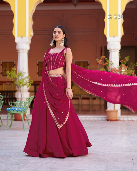 A woman models a deep magenta lehenga choli set with gold embroidery, standing in front of a traditional courtyard with yellow arches. The dupatta is spread out to the side, showing its gold border and small gold embellishments.
