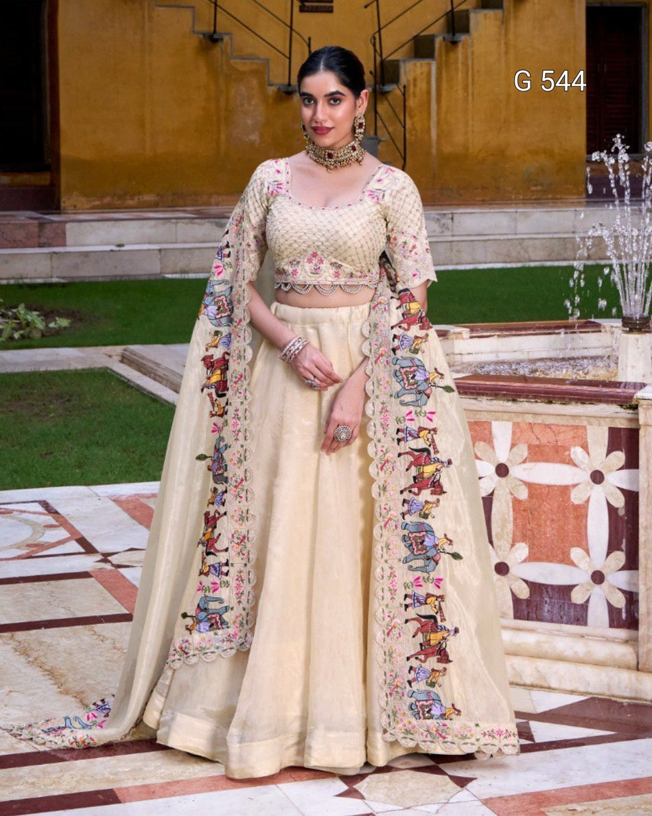A woman models a traditional Indian ethnic outfit consisting of a cream-colored flared skirt (lehenga), a matching embroidered blouse, and a sheer embroidered dupatta, set against an outdoor architectural background with a fountain.