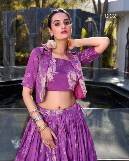 A woman modeling a purple and white printed traditional outfit consisting of a crop top, short jacket, and flared skirt, with gold jewelry and a flower earring, set against an outdoor architectural background.