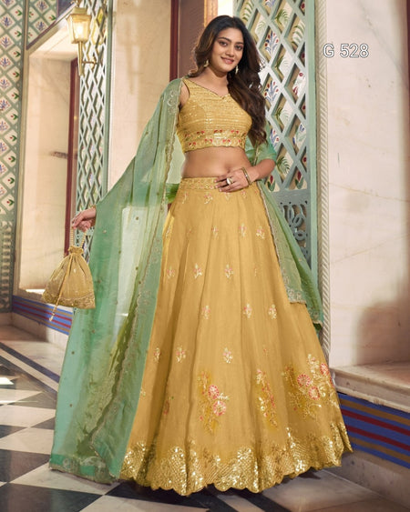 A woman models a traditional Indian lehenga choli set in a decorative architectural corridor. The outfit consists of a mustard yellow embroidered skirt (lehenga), a matching embroidered crop top (choli), and a sheer mint green dupatta draped over her arm and held in one hand.