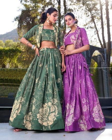 Two women modeling traditional Indian lehenga choli outfits outdoors. The woman on the left wears a green floral print outfit with a matching jacket, and the woman on the right wears a purple floral print outfit with a matching jacket.