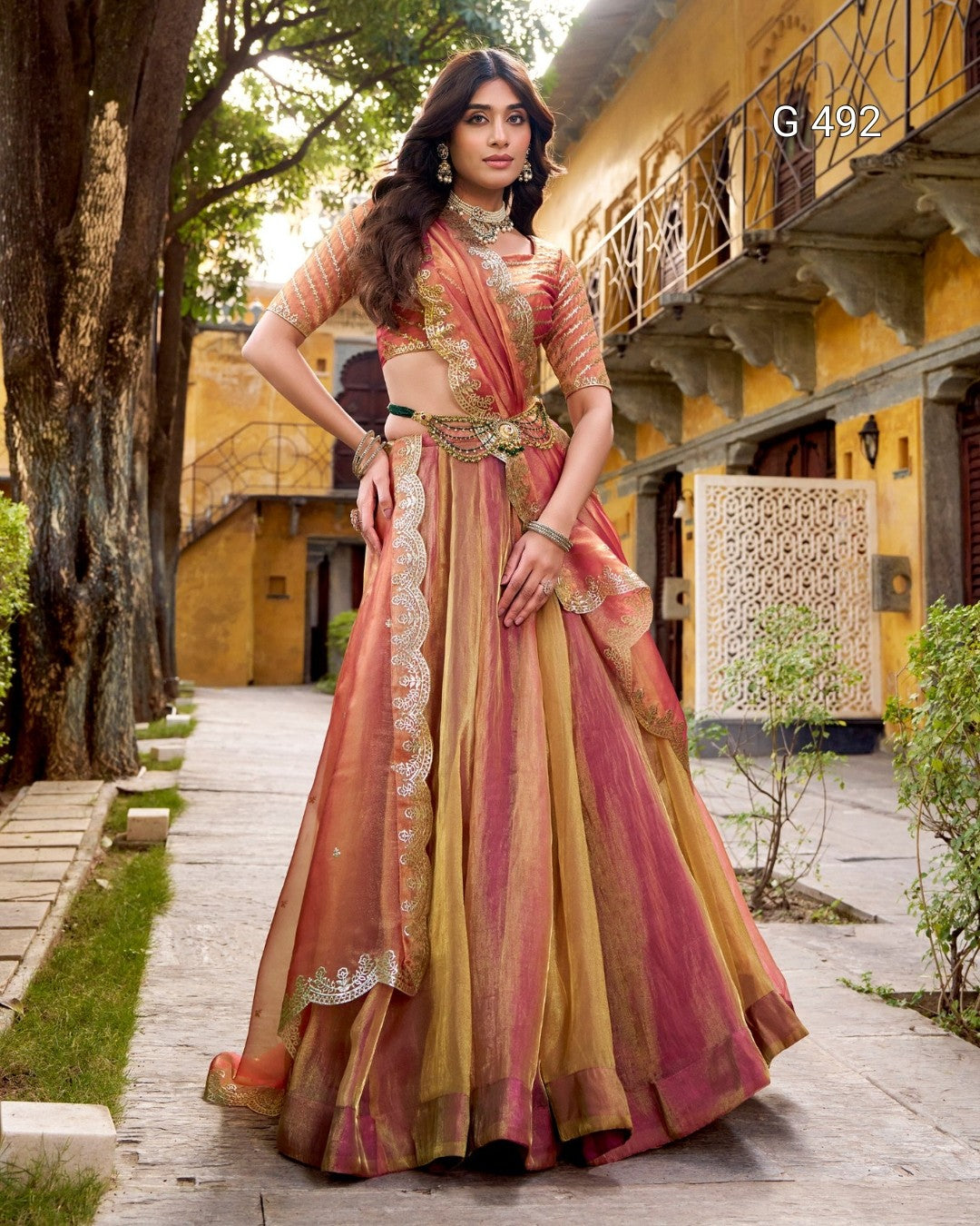 A woman models a traditional Indian ethnic outfit consisting of a peach, pink, and gold printed skirt with a matching blouse, standing on a stone pathway in front of a yellow building.