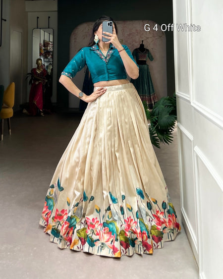 A woman takes a mirror selfie wearing a traditional Indian lehenga choli set. The set features a teal green embroidered blouse and a voluminous off-white skirt with a vibrant floral border print.