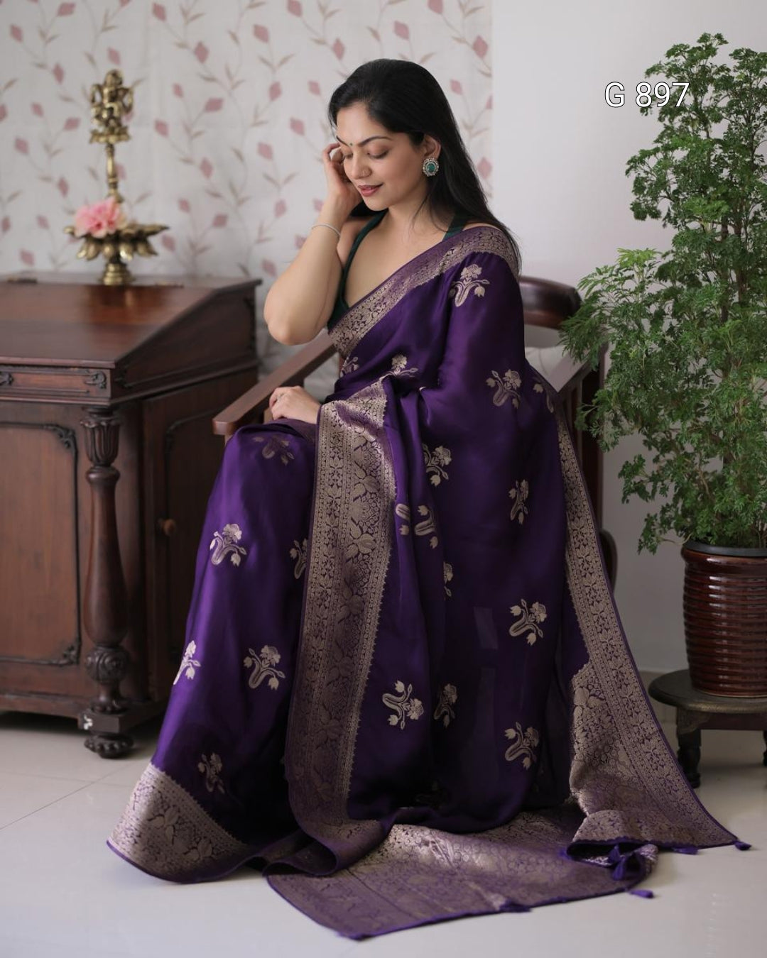 Woman wearing a purple saree with gold patterns, sitting indoors.