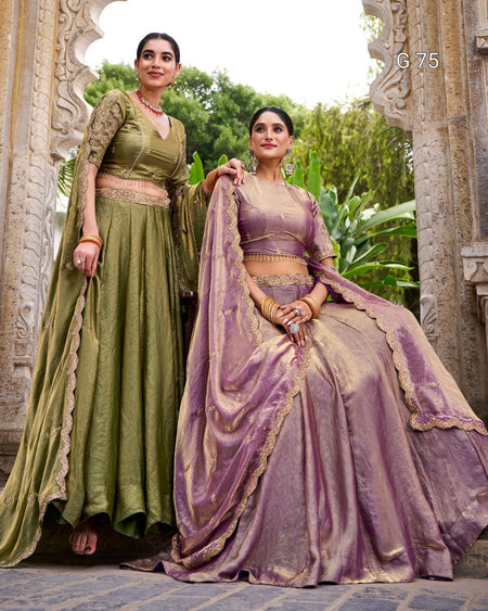 Two women modeling traditional Indian ethnic wear in an outdoor stone archway setting. One wears an olive green anarkali-style dress with beige lace trim, and the other wears a lavender-purple lehenga choli set with gold zari detailing and a matching dupatta.