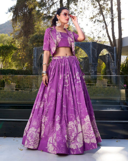 A woman models a purple and beige floral printed Indian ethnic wear set consisting of a kurta, matching crop jacket, and a full flared skirt, set against an outdoor background with trees and a stone structure.