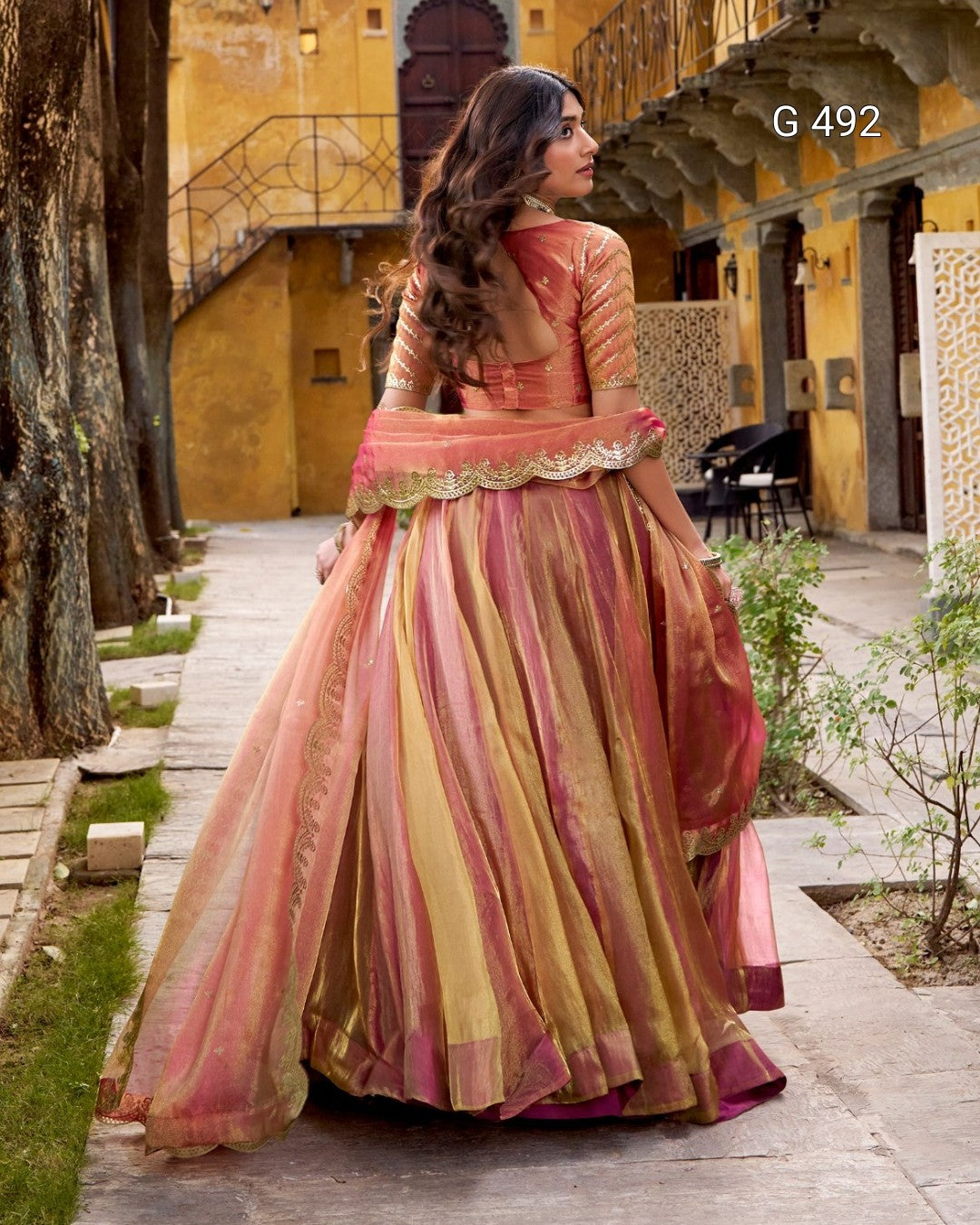 A woman wearing a traditional Indian salwar kameez outfit with a pink and gold striped skirt, standing in a courtyard with yellow walls.
