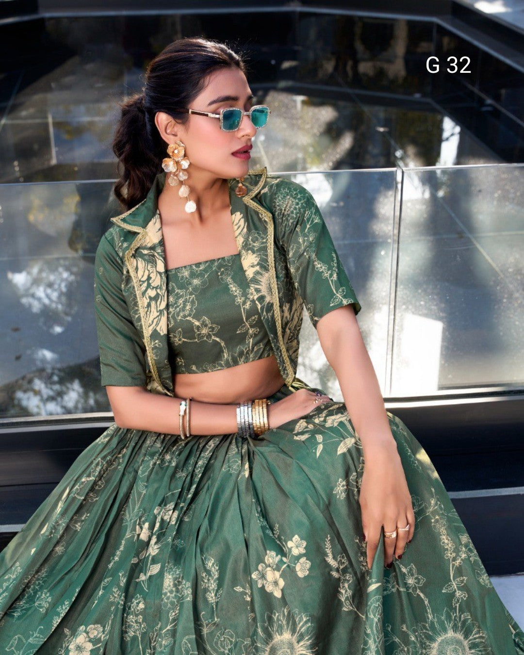 A woman models a green floral print lehenga choli set with a matching jacket, seated in front of a glass car window. The outfit features a square-necked choli, a full flared skirt, and gold-toned floral embroidery and print details.