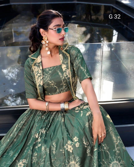 A woman models a green floral print lehenga choli set with a matching jacket, seated in front of a glass car window. The outfit features a square-necked choli, a full flared skirt, and gold-toned floral embroidery and print details.