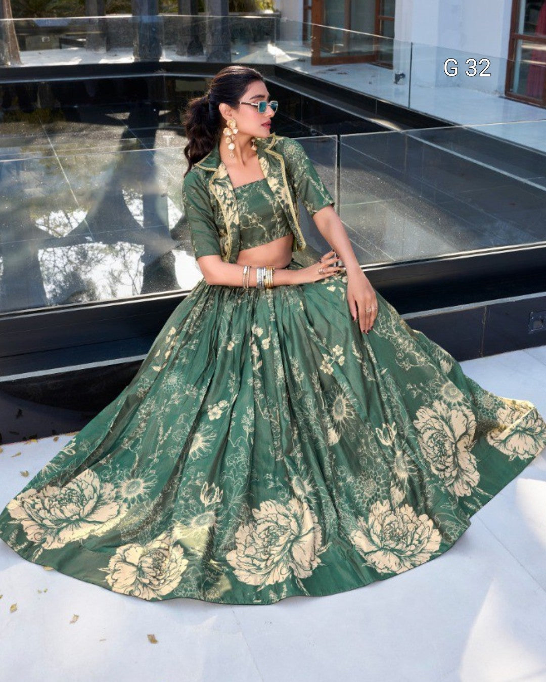 A woman models a green and gold floral print lehenga choli set in front of a modern glass pool. The set features a green printed blouse with short sleeves and a matching full flared skirt with large gold-toned floral motifs.