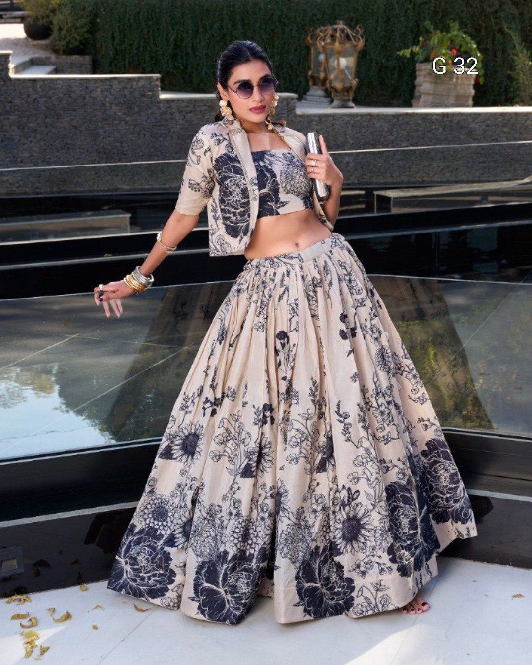 A woman models a beige and navy floral printed traditional outfit consisting of a cropped jacket, crop top, and a voluminous flared skirt, set against a modern glass and stone terrace background.