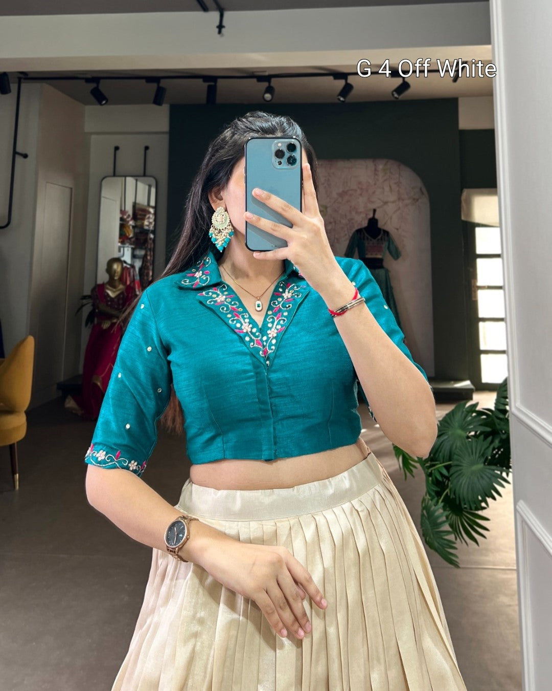 A woman takes a mirror selfie in a boutique wearing a teal short-sleeve crop top with colorful embroidered detailing on the collar and cuffs, paired with a pleated cream skirt. The text 'G 4 Off-White' is visible in the top right corner.