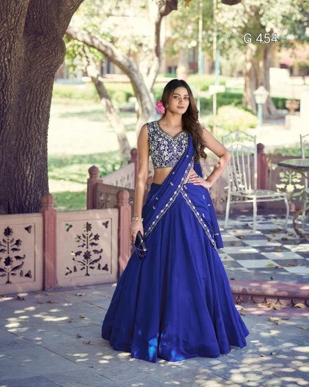 A woman stands outdoors wearing a royal blue lehenga choli with silver embroidery. The scene features a stone pathway, a large tree trunk, and garden chairs in the background.