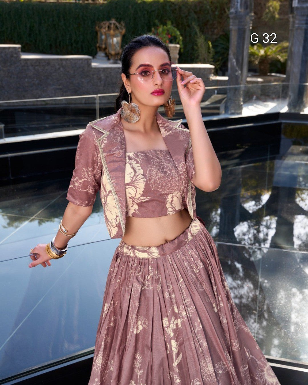 A woman models a dusty rose and gold printed traditional outfit consisting of a crop top, short jacket, and pleated skirt, standing in front of a reflective glass pool.