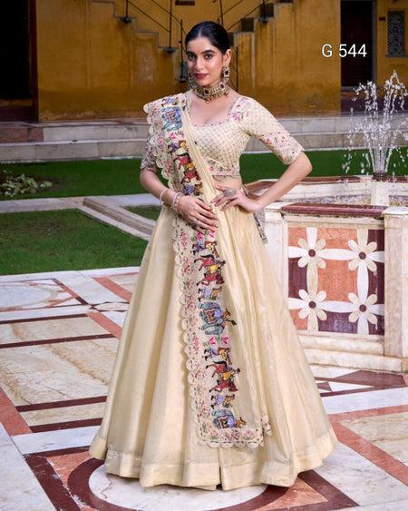 A woman models a traditional Indian ethnic outfit consisting of a beige flared gown (lehenga), a matching embroidered blouse, and a draped dupatta, set against an outdoor courtyard background with a fountain.