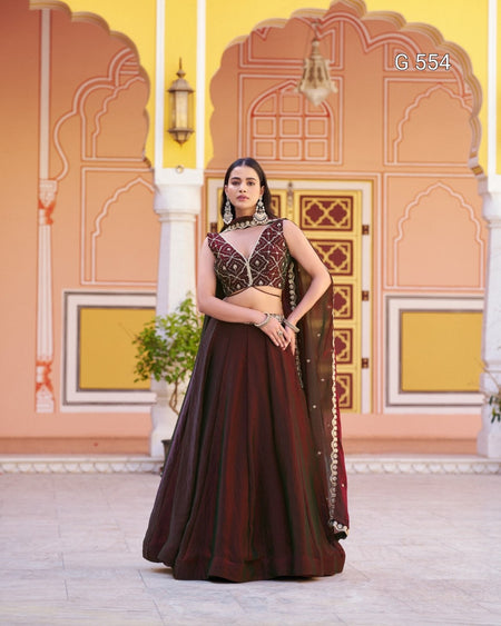 A woman models a traditional Indian lehenga choli dress in deep maroon with intricate gold embroidery, standing in front of a pink and yellow architectural backdrop.