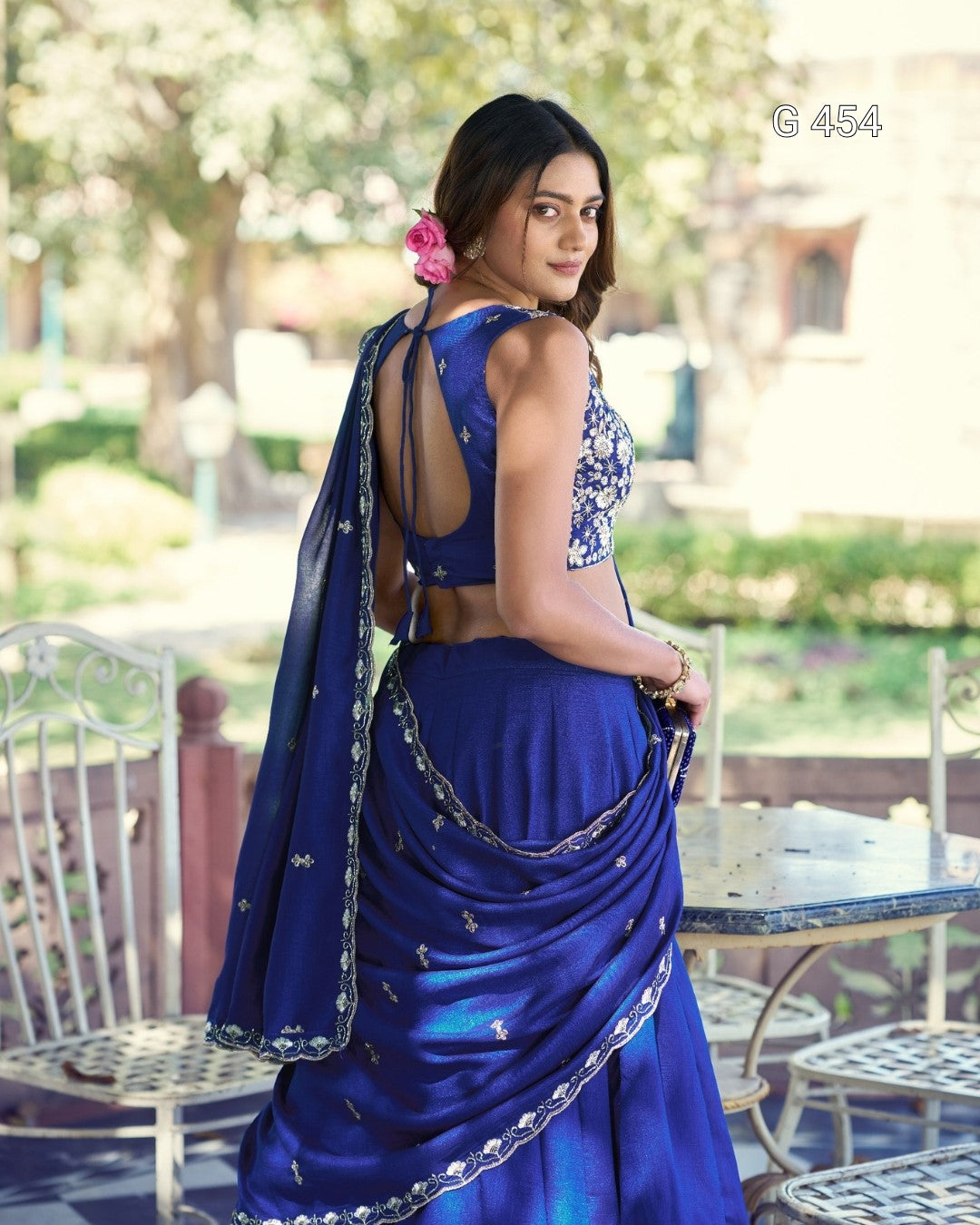 A woman wearing a royal blue traditional Indian saree with silver embroidery and a matching blouse, photographed outdoors on a patio with garden furniture and blurred trees in the background.