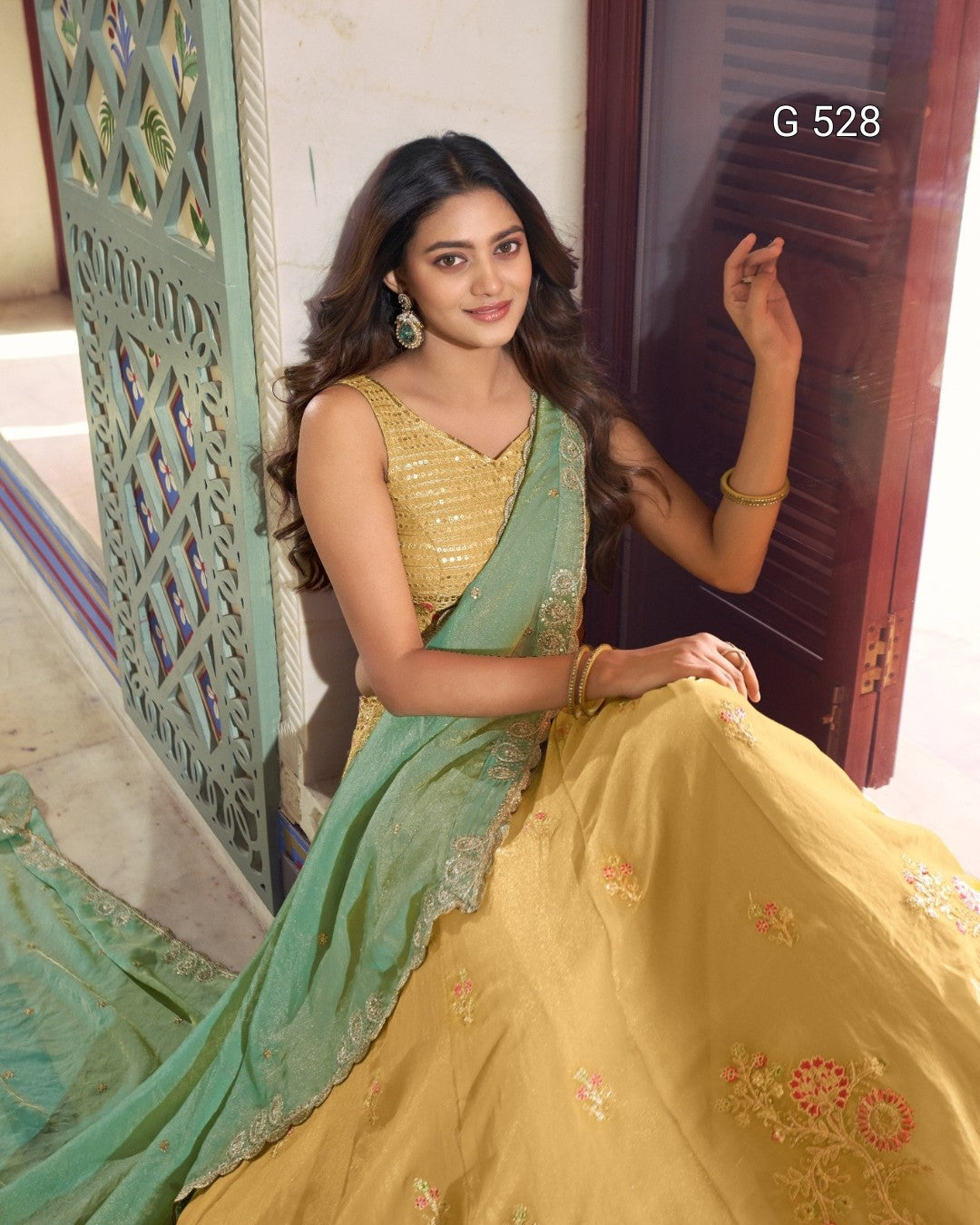 A woman wearing a traditional Indian outfit with a mustard yellow embroidered skirt and a mint green dupatta, posed indoors next to a carved screen and wooden door. The text 'G 528' is visible in the top right corner.