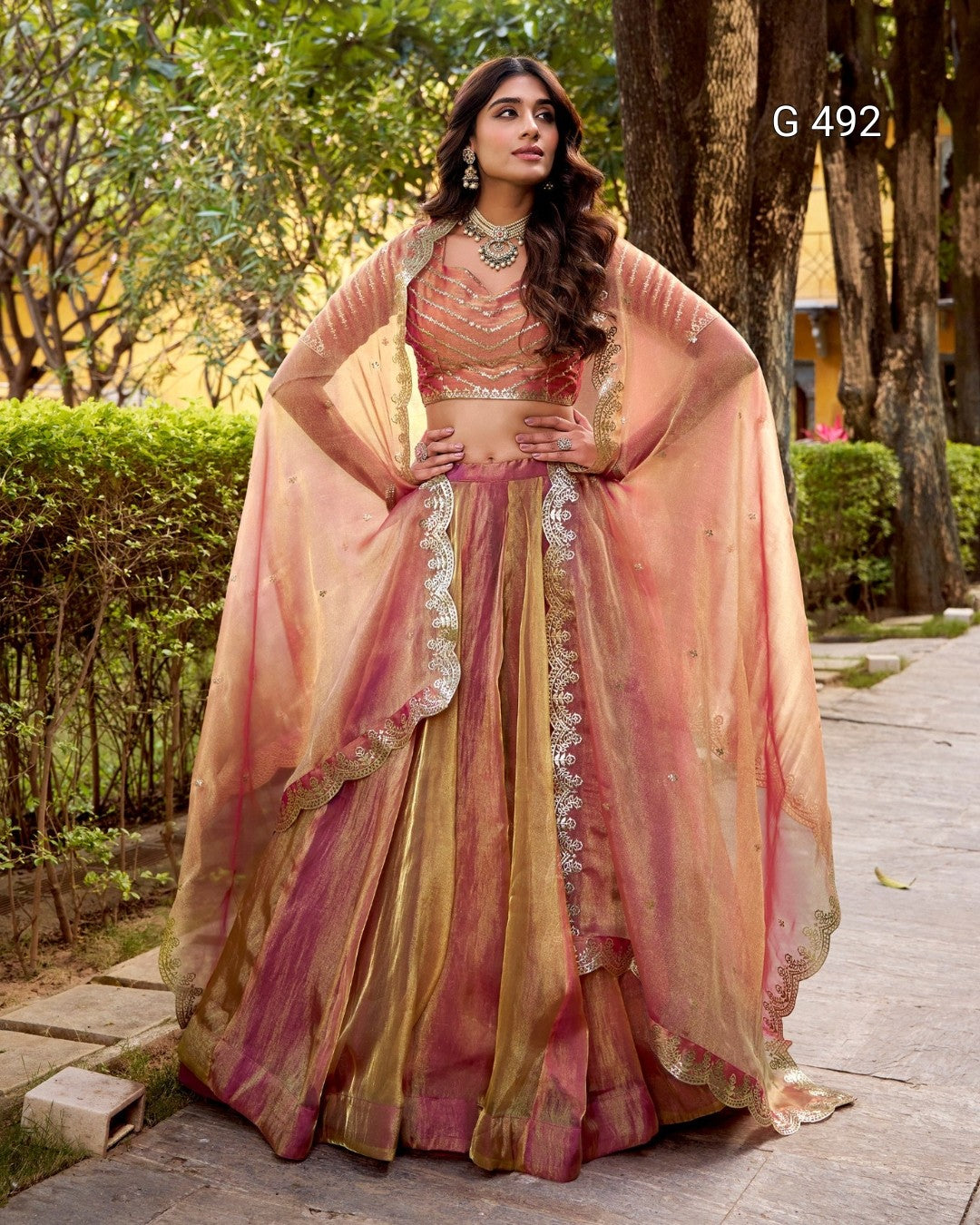 A woman stands outdoors on a paved walkway wearing an ornate peach-pink and gold traditional outfit with a sheer draped overlay and embroidered details. Trees and hedges are visible in the background, and the text 'G 492' appears near the upper right.