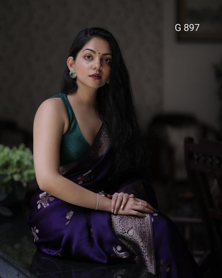 Woman in a green and purple saree sitting indoors with a blurred background