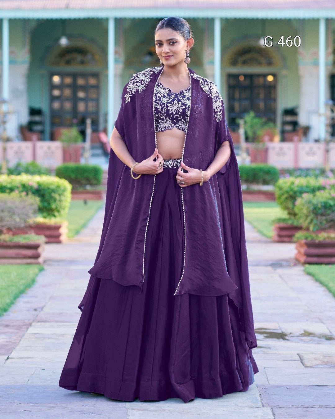 A woman stands outdoors wearing a traditional Indian outfit consisting of a deep purple skirt, a matching embroidered blouse, and a draped purple shawl. The background shows a garden path and a building with columns.