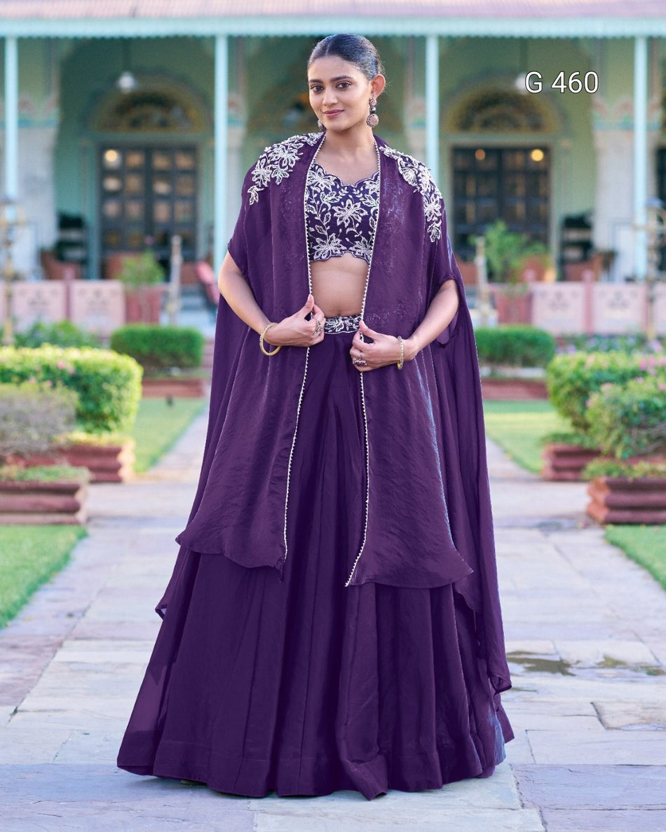 A woman stands outdoors wearing a traditional Indian outfit consisting of a deep purple skirt, a matching embroidered blouse, and a draped purple shawl. The background shows a garden path and a building with columns.