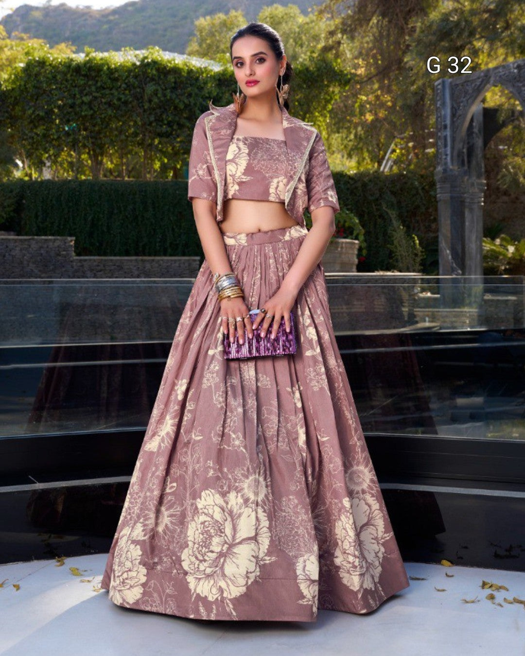 A woman stands outdoors wearing a dusty mauve lehenga choli set with beige floral motifs. The set includes a short-sleeve crop top, matching high-waisted skirt, and a small matching clutch bag.
