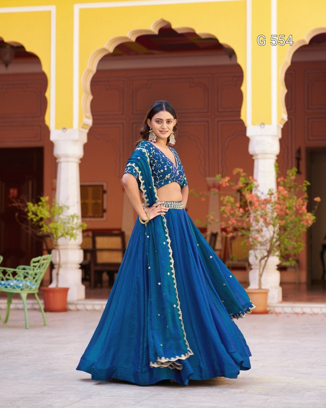 A woman models a traditional Indian lehenga choli set in a courtyard with yellow walls and arches. The outfit is a vibrant teal blue with gold embroidery and border details.