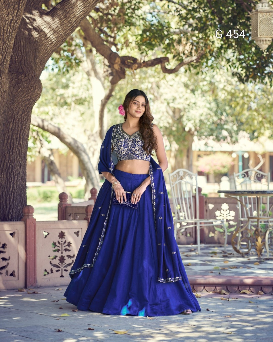 A woman stands outdoors under a tree wearing a royal blue traditional outfit with a heavily embroidered silver and gold blouse, matching skirt, and dupatta. The setting includes patio furniture and a decorative wall in the background.