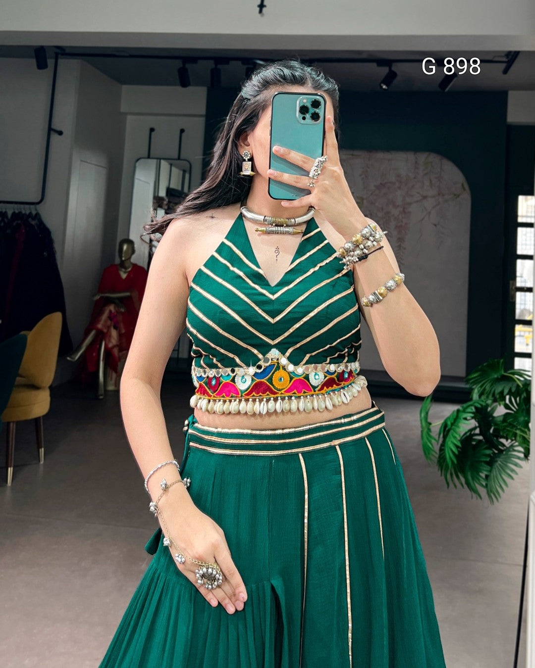 Woman wearing a green traditional outfit with intricate designs, holding a phone in a room.