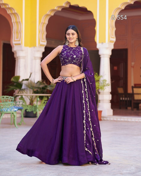 A woman models a traditional Indian outfit consisting of a purple flared skirt (lehenga), a matching embroidered crop top (choli), and a draped scarf (dupatta) with gold embroidery, set against a background of yellow and white arches.