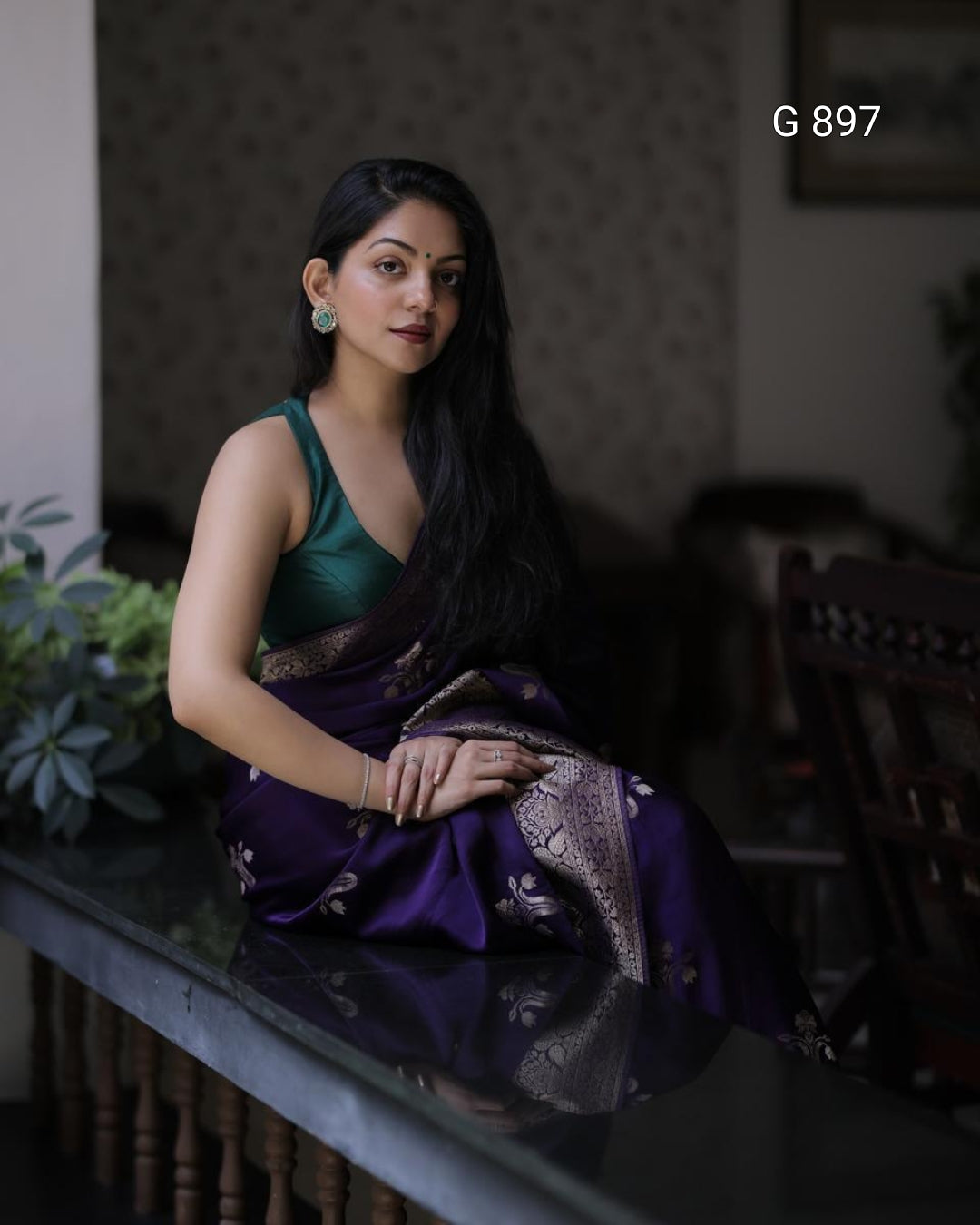 Woman in a green and purple saree sitting indoors with a blurred background