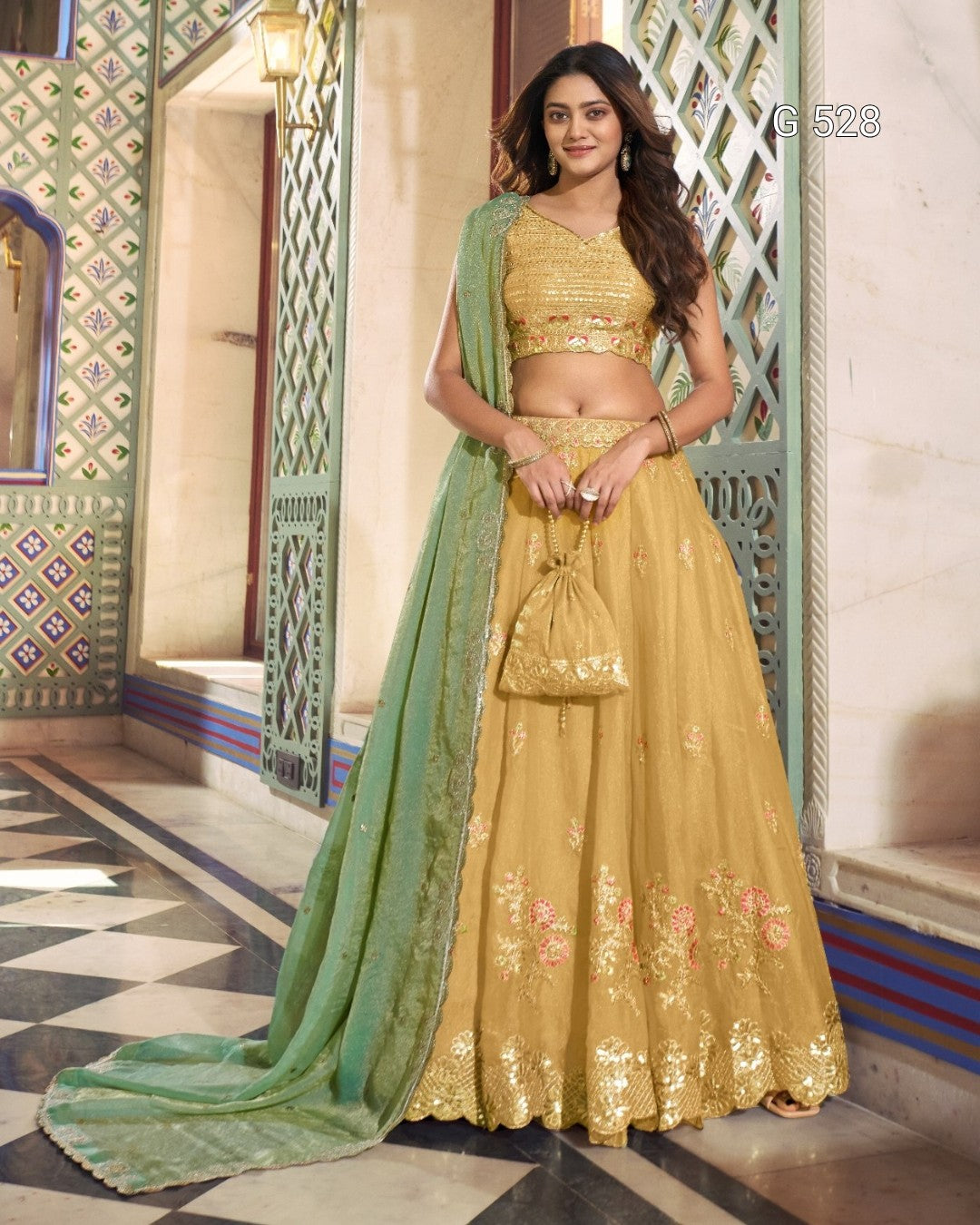 A woman models a traditional Indian lehenga choli set in a decorative architectural corridor. The outfit consists of a mustard yellow embroidered skirt (lehenga), a matching embroidered crop top (choli), and a sage green dupatta draped over her shoulder.