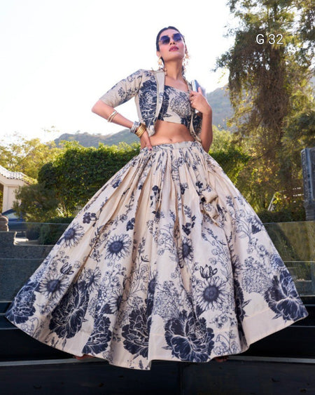 A woman wearing a beige and navy floral printed lehenga choli set with a matching jacket, standing outdoors on stone steps with trees and hills in the background.