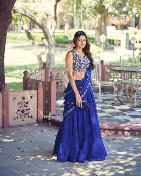 A woman stands outdoors in a park-like setting wearing a royal blue traditional outfit with a heavily embellished silver sleeveless top and a matching flared skirt with silver border detailing. She is holding a small metallic clutch.
