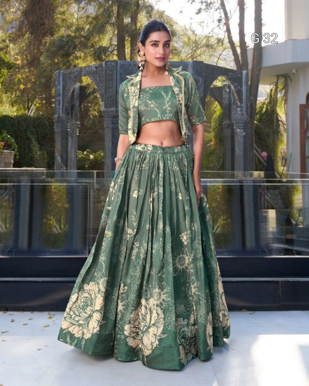 A woman models a green two-piece traditional outfit with a matching printed jacket, featuring a high-waisted flared skirt and a cropped jacket, set against an outdoor architectural background.