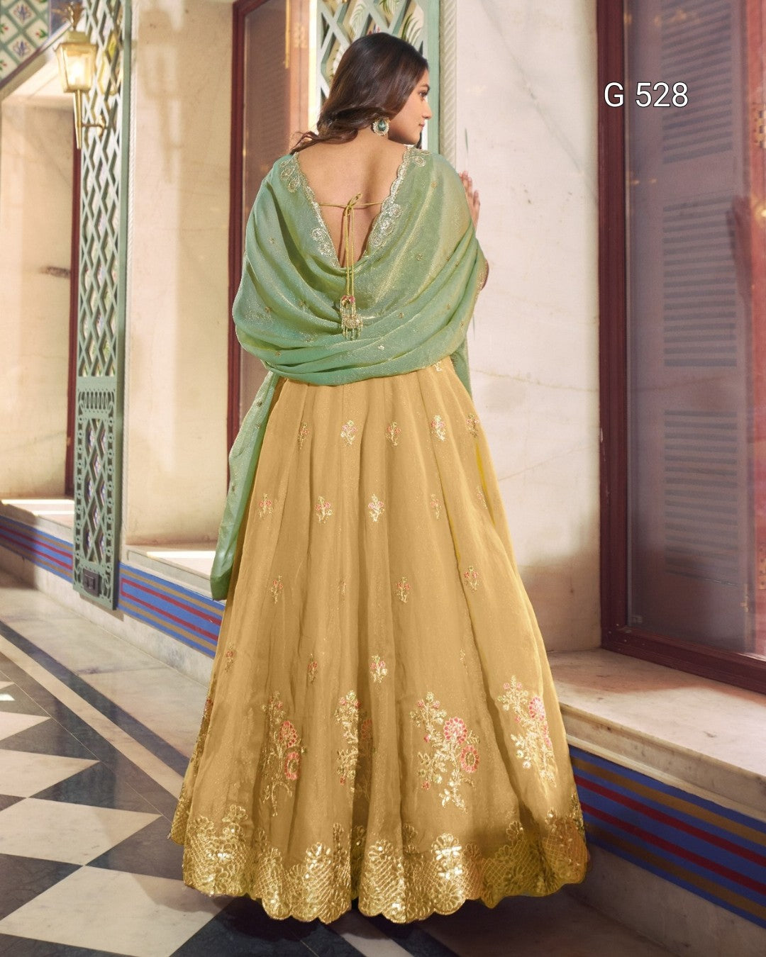 A woman shown from the back wearing a traditional Indian outfit consisting of a mustard yellow embroidered skirt (lehenga), a sage green dupatta, and a matching green embroidered blouse. The setting is a traditional courtyard with stone walls and tiled floors.