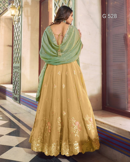 A woman shown from the back wearing a traditional Indian outfit consisting of a mustard yellow embroidered skirt (lehenga), a sage green dupatta, and a matching green embroidered blouse. The setting is a traditional courtyard with stone walls and tiled floors.